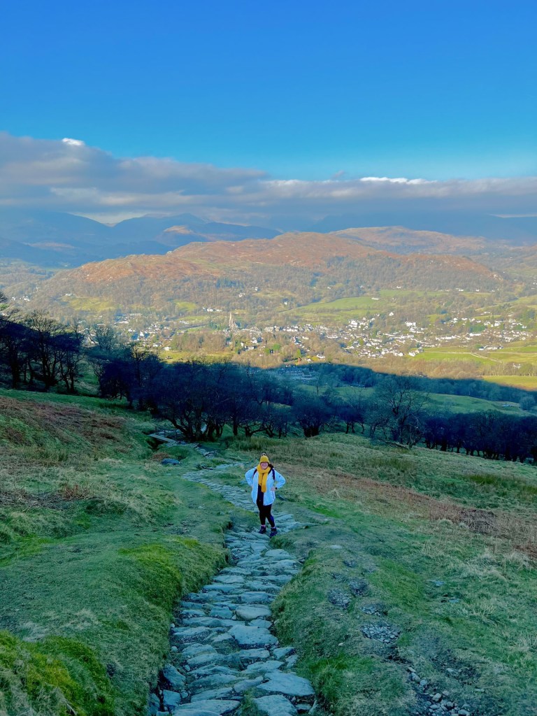 A photo looking down the long stone path that leads up to Wansfell. April stood half-way looking up with her hands on her hips and smiling. In the background in the town of Ambleside and fells in the distance. Bright blue skies with clouds on the horizon.