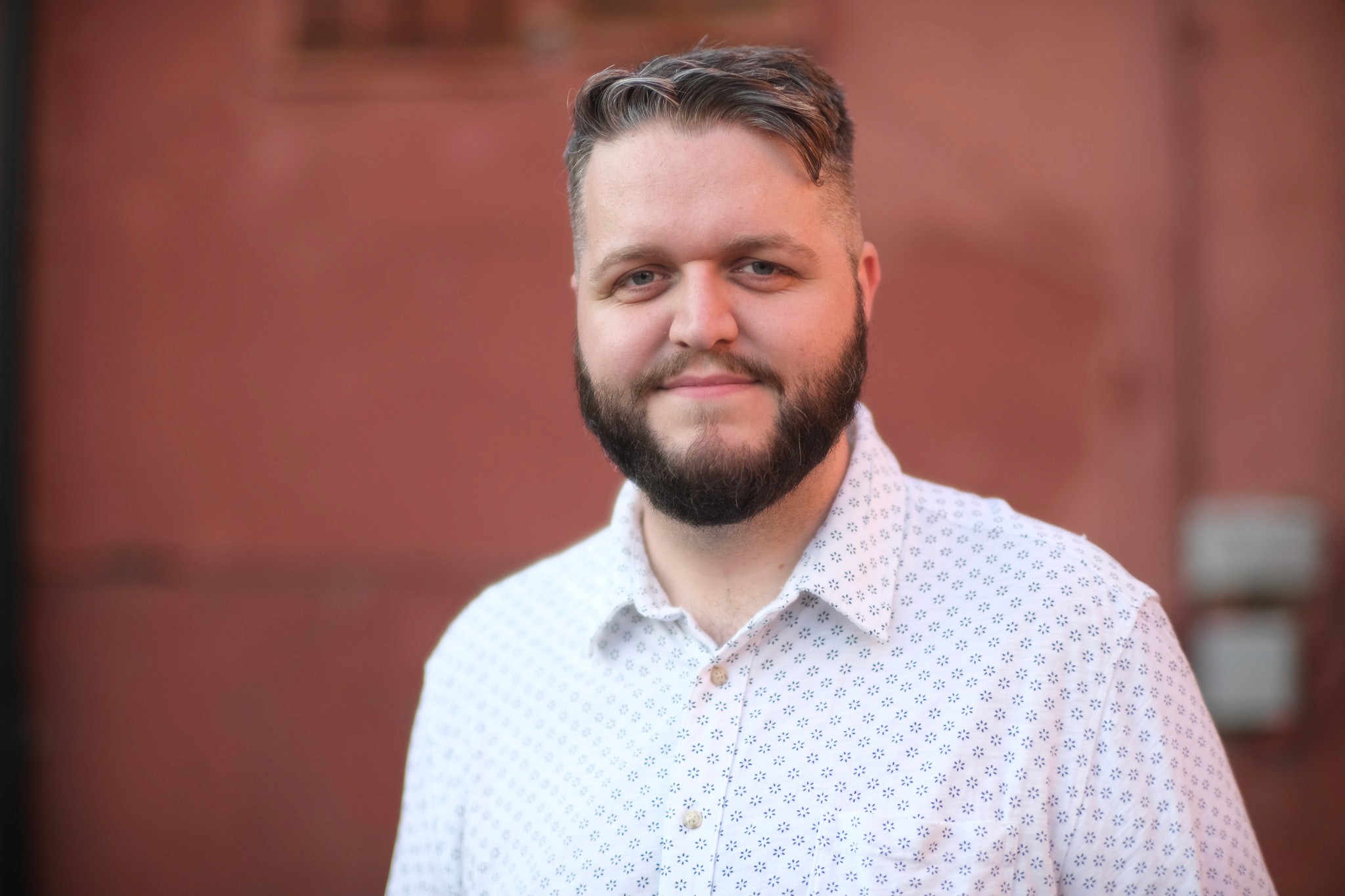 Portrait of Will Peterson, wearing a light-colored button-up shirt with a small dark pattern. He is standing in front of a reddish wall and smiling slightly at the camera.