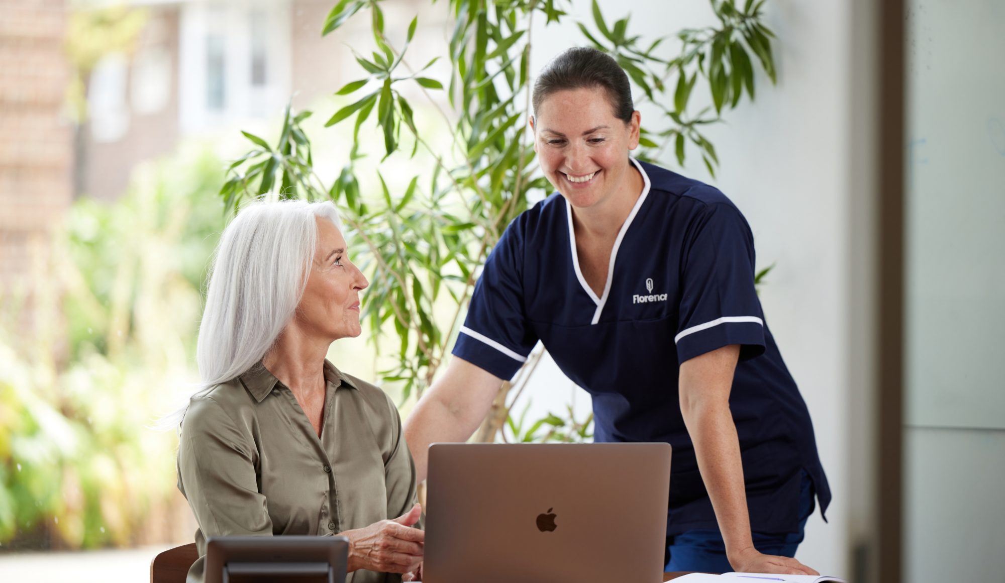 A smiling Florence nurse in navy uniform standing beside an older woman seated at a desk with a laptop, both engaged in a warm interaction