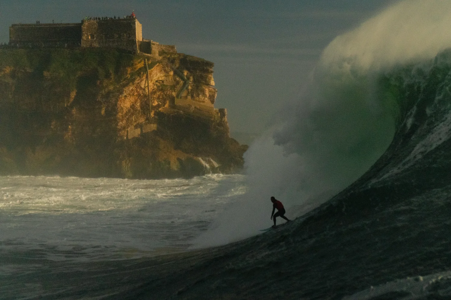 Surfer riding a huge wave in Nazaré, with the cliff and fort in the background.