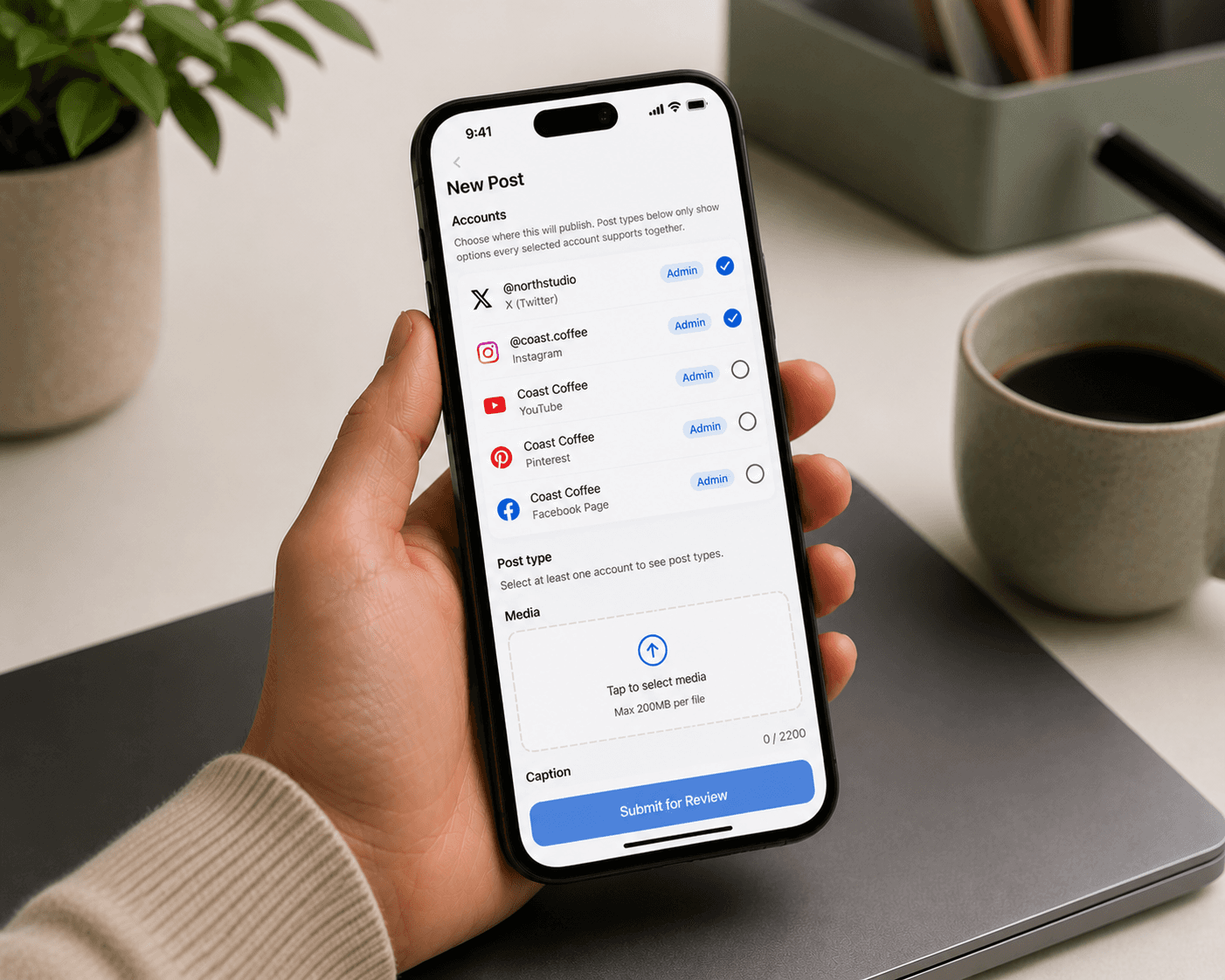 A person holds a smartphone displaying a social media management app interface with account options and post creation settings, set against a cozy workspace background featuring a ceramic mug and a potted plant.