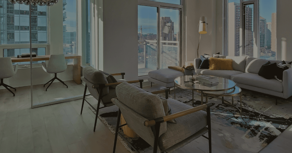 A condominium living room with large bright windows, two grey simple arm chairs with grey upholstery and wooden arm rests. Across the room is a large low sofa with thin legs. The centre has two circle glass tables. The room is finished with an area rug. This is an example of scandinavian design.