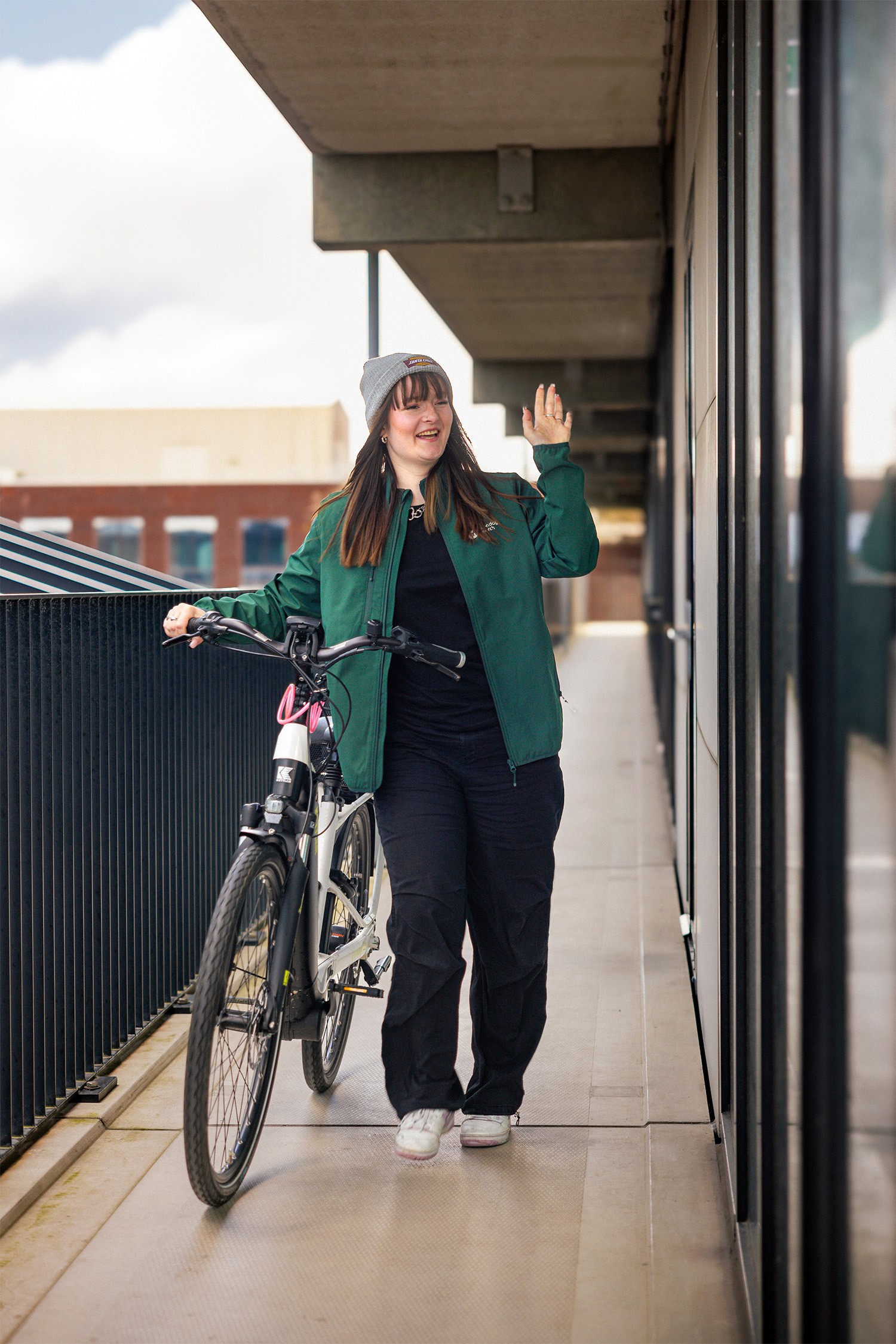 A person in a green jacket walks a bicycle, smiling and waving