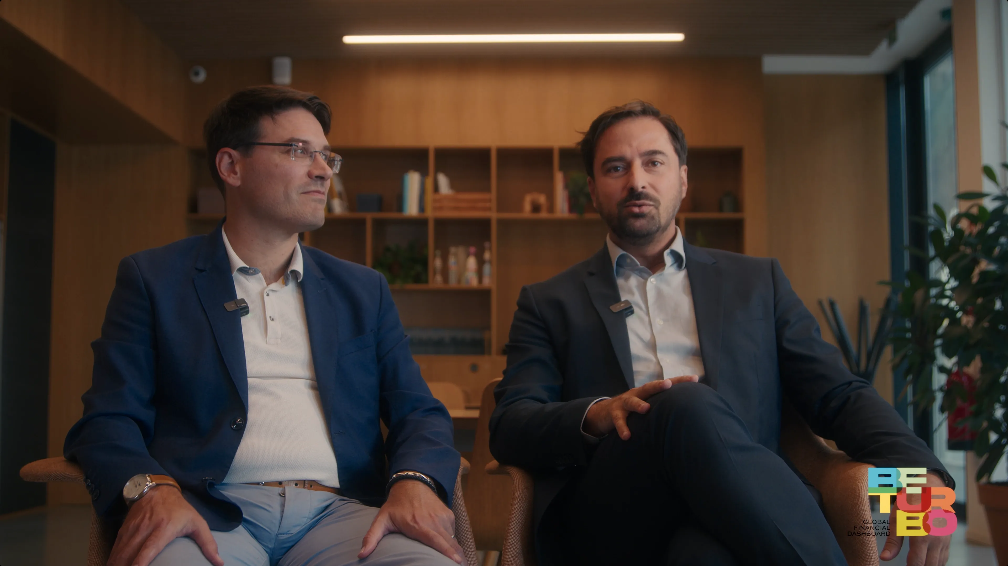 Two men in suits are seated in a modern office setting, engaged in a discussion with books and plants visible in the background.