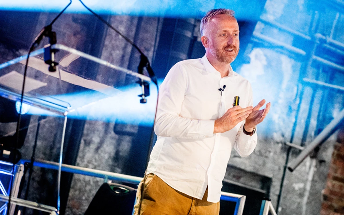 A person in a white shirt stands on a stage, speaking and gesturing with their hands, with bright blue lighting and industrial-style staging in the background.