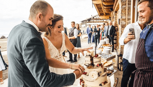 Brautpaar schneidet gemeinsam die Hochzeitstorte während Hochzeitsfeier mit Gästen im Hintergrund