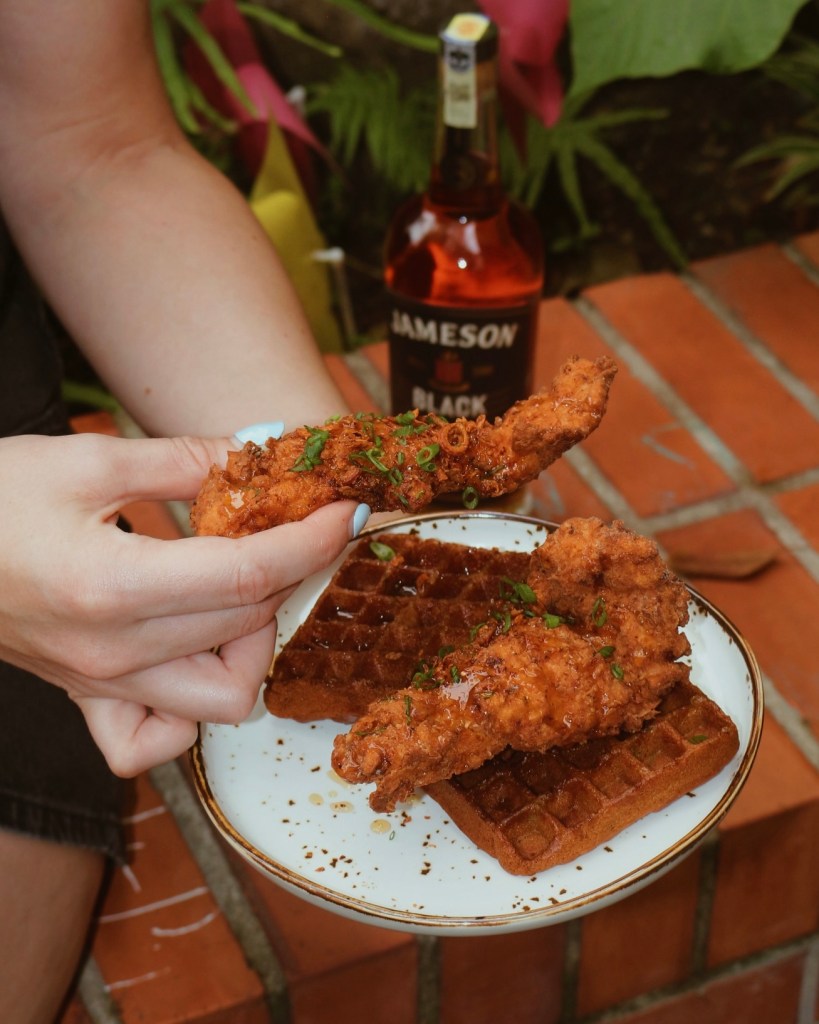 A person's hand holding a piece of fried chicken above a plate with waffles, drizzled with syrup, with a bottle of Jameson Black Barrel whiskey in the background.