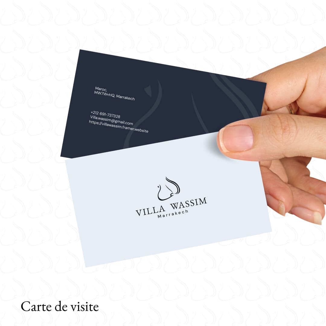 {  Mise en situation (mockup) d'une carte de visite pour 'Villa Wassim Marrakech'. La carte, tenue par une main, présente un design double face : le recto blanc affiche le logo élégant (silhouette de cheval ou arabesque) et le nom de la marque, tandis que le verso bleu nuit contient les coordonnées (téléphone, email, site web, adresse) dans une typographie minimaliste. Ce visuel démontre l'expertise de Nux-Studio en design graphique, conception de papeterie de luxe et maintien de la cohérence de l'identité visuelle de marque (branding) sur tous les supports imprimés.