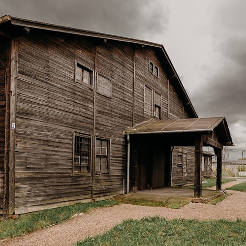 Weathered wooden building with boarded-up windows, a covered entrance, gravel pathway, and a cloudy sky overhead.
