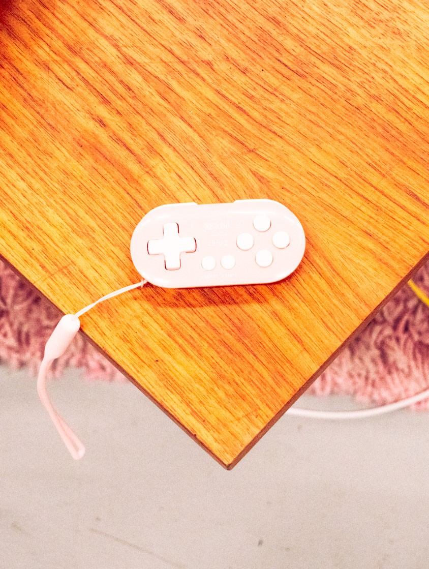 A pastel pink game controller with a wrist strap sits on a wooden surface.