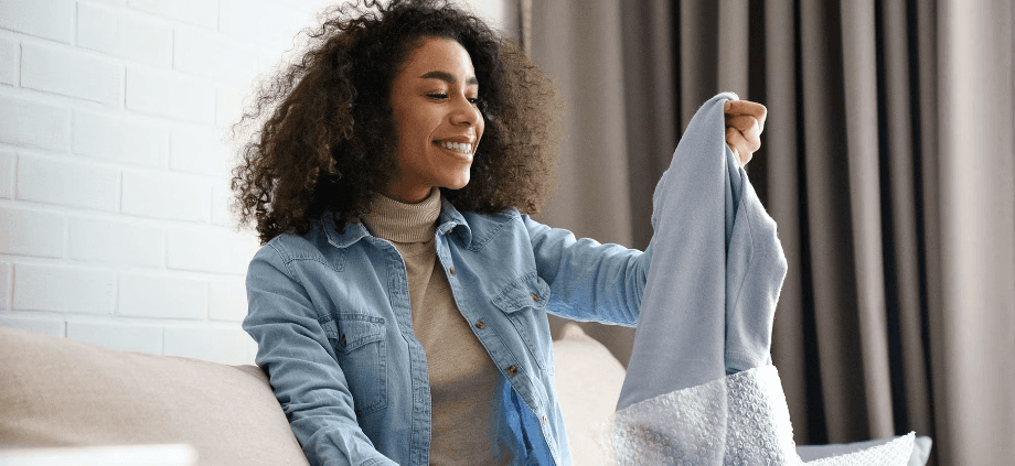 a smiling woman lifts a light blue sweater out of a shipping box