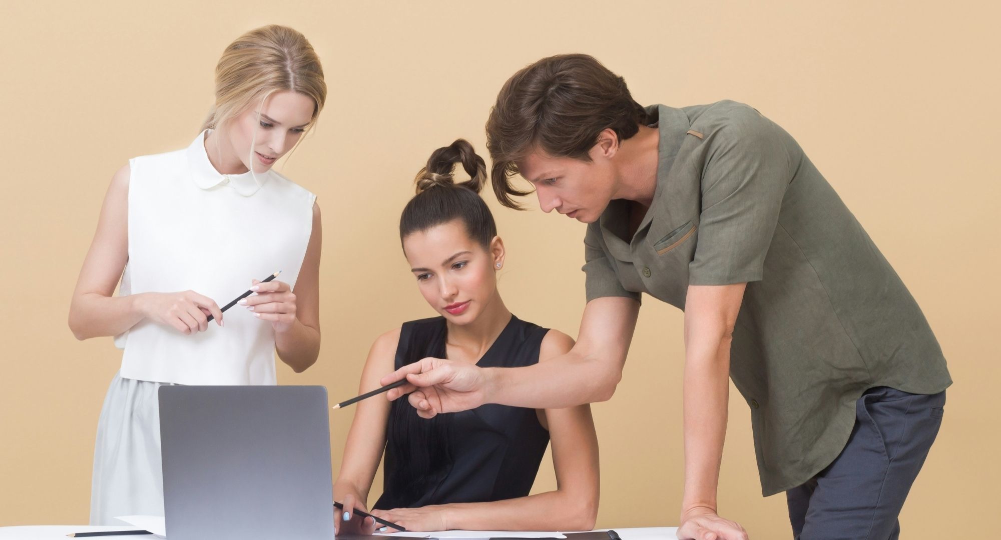 três pessoas (duas mulheres e um homem) numa sala de parede cor bege olhando para a tela de um notebook, a mulher do meio vestida de preto está sentada, a outra mulher, vestida de branco, está à sua direita e o homem a sua esquerda, apontando com um lápis para a tela