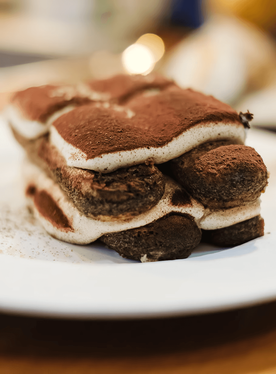 A close-up of a slice of tiramisu with layers of cocoa-dusted sponge and creamy mascarpone, served on a white plate with a softly blurred background.