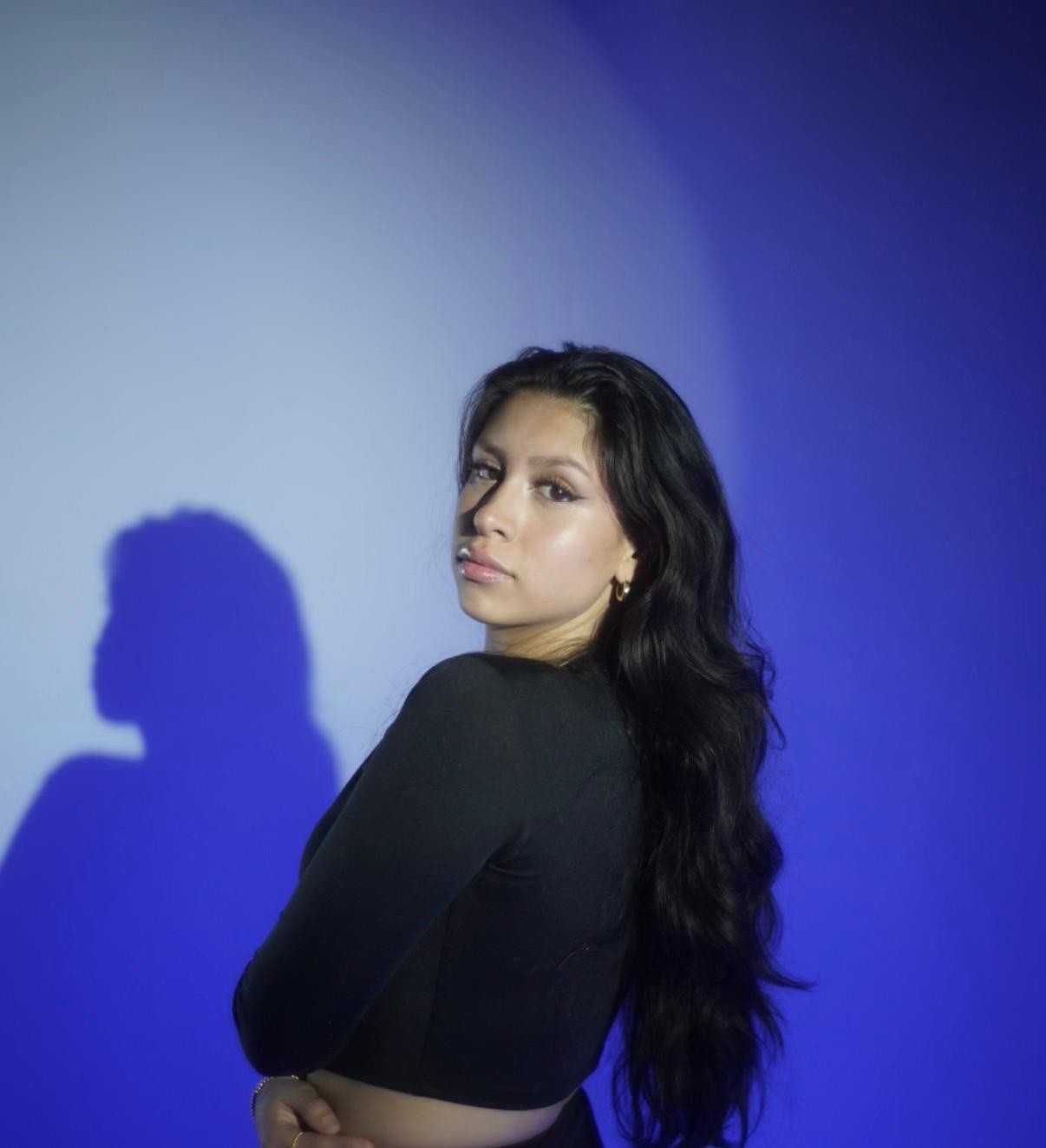 A person with long dark hair and wearing a white top holds a shiny silver purse, standing in a modern, dimly lit room decorated with various photographs on the walls.
