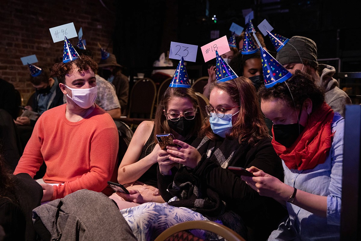 A cluster of audience members wearing. party hats gather around to look at someone's phone.