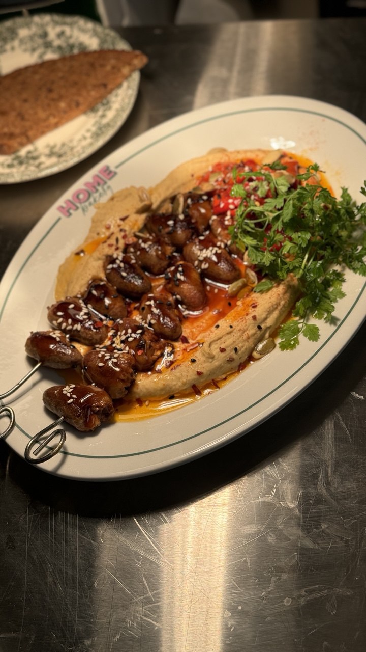 A dish of grilled meat skewers is garnished with sesame seeds and served on a white oval platter with creamy hummus, a splash of red sauce, and a fresh cilantro garnish, placed atop a stainless steel surface.
