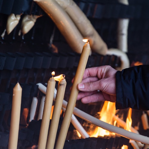 يد تمسك بشمعة مضاءة تضيء شموعًا أخرى أمام رف مليء بالشموع المذابة جزئيًا، مع ألسنة اللهب في الخلفية.