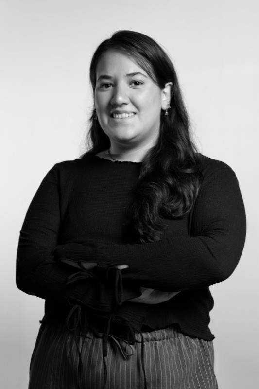 Black-and-white studio portrait of a smiling woman with long dark hair, wearing earrings, a necklace, and a black knit top with sleeves tied at the cuffs, arms crossed, against a plain light background.