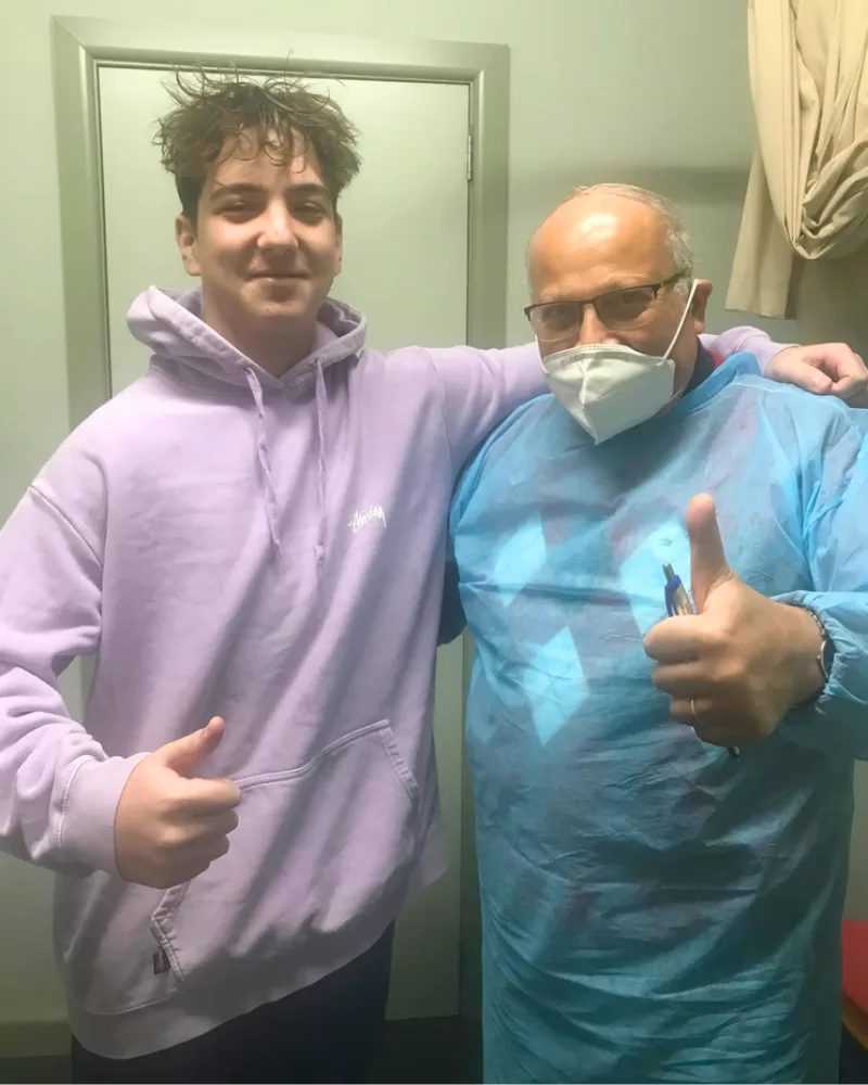 Dr. Ashraf Saddik giving a thumbs-up beside a young adult patient in a purple hoodie inside a Coburg clinic room.