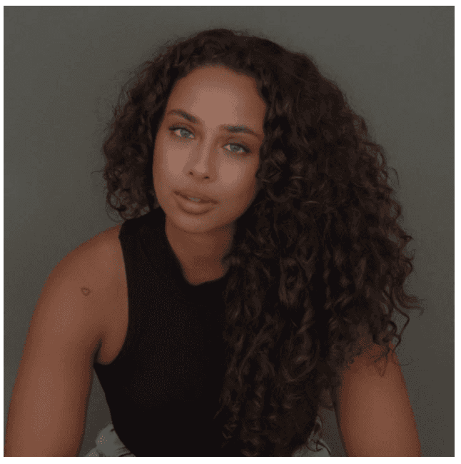 Portrait of a young man with wavy dark hair and a trimmed beard in a white sweater, soft light on a neutral background.