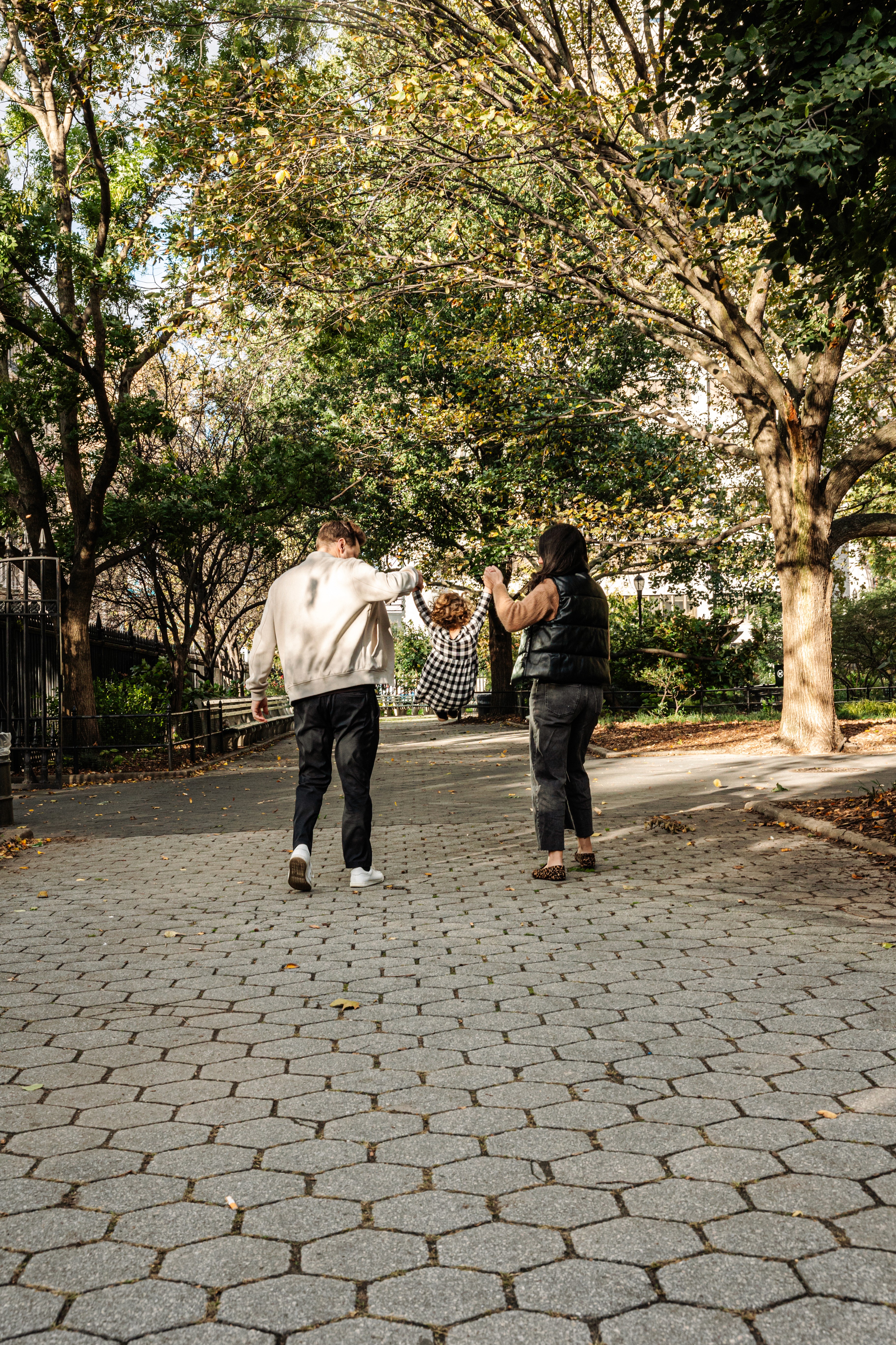5:54 PMMom and dad each holding their daughter's hand, swinging her through the air mid-stride at Stuyvesant Square Park in the Gramercy and East Village neighborhood of NYC — joyful, candid family photography by Lizz Spano Photography, New York City family photographer.