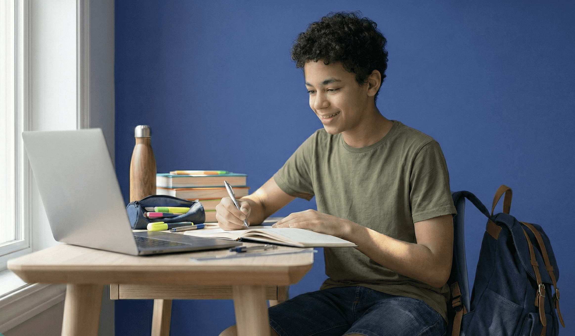 High school student studying at a desk with a laptop and notebook, taking notes in a home study environment.