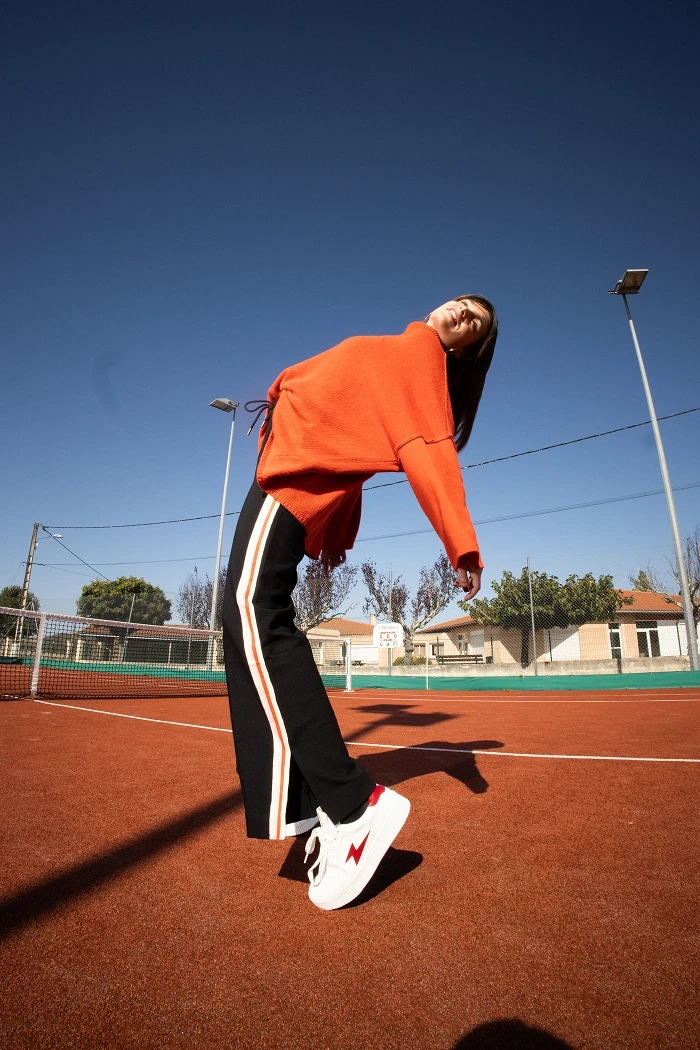 Une femme avec un pull orange, pose sur un court de tennis.
