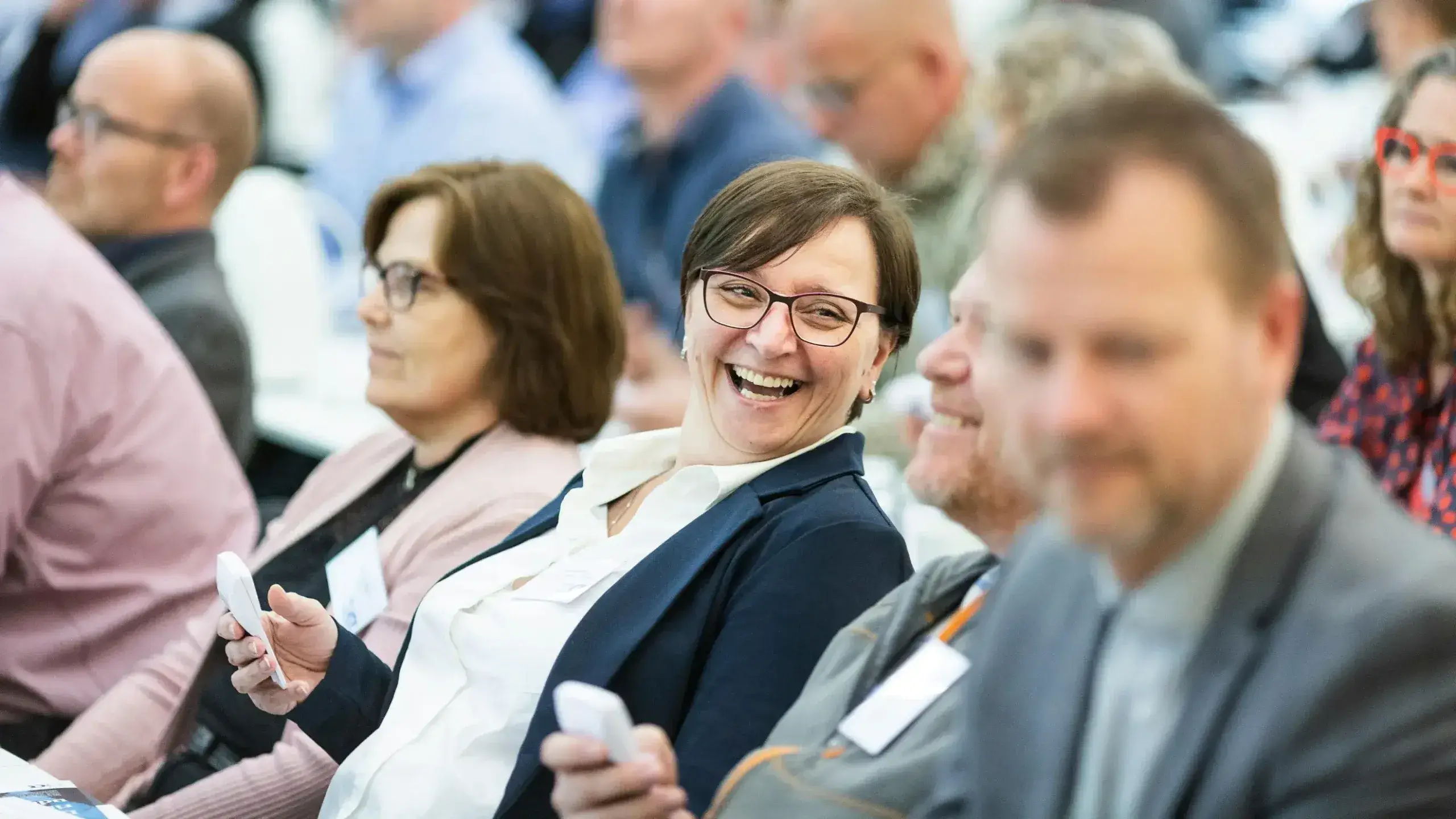 A woman in business attire smiles and holds an electronic device among a seated group at a conference, creating a lively, engaged atmosphere.