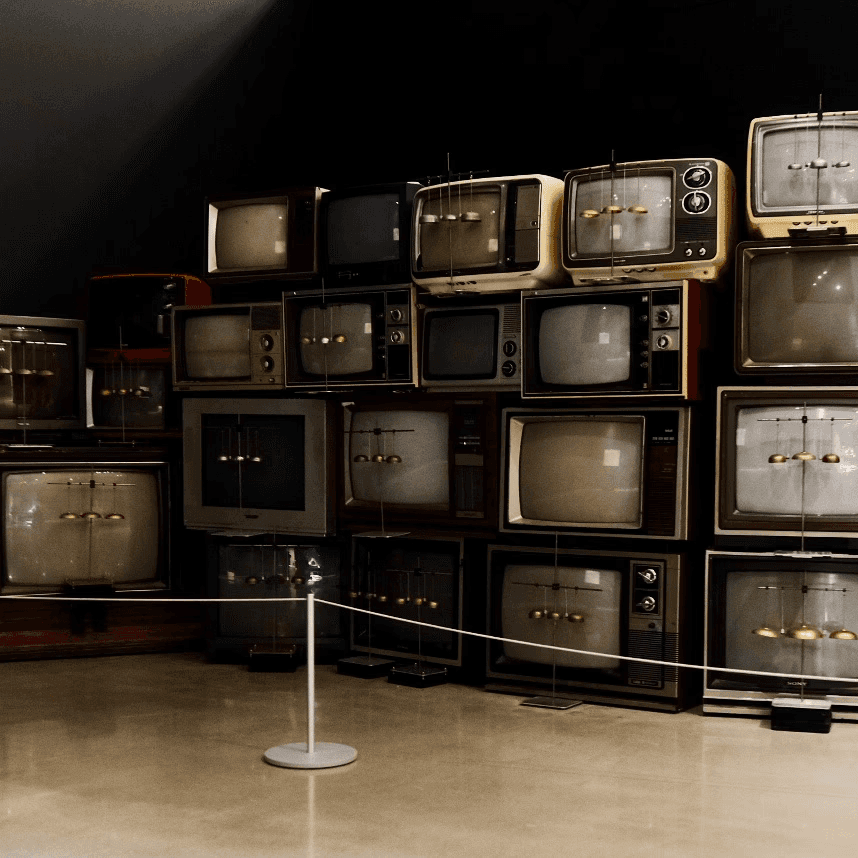 A museum installation titled 'Electrostatic Bell Choir' featuring a large wall of stacked vintage cathode-ray tube televisions from the mid-20th century. Each TV screen displays a small suspended pendulum or bell mechanism, suggesting the sets generate electrostatic fields to animate the bells. The televisions vary in size, color, and style — ranging from cream and yellow to dark wood-toned models — and are arranged in a roughly pyramidal formation against a dark wall. A rope barrier in the foreground indicates this is a gallery exhibit.
