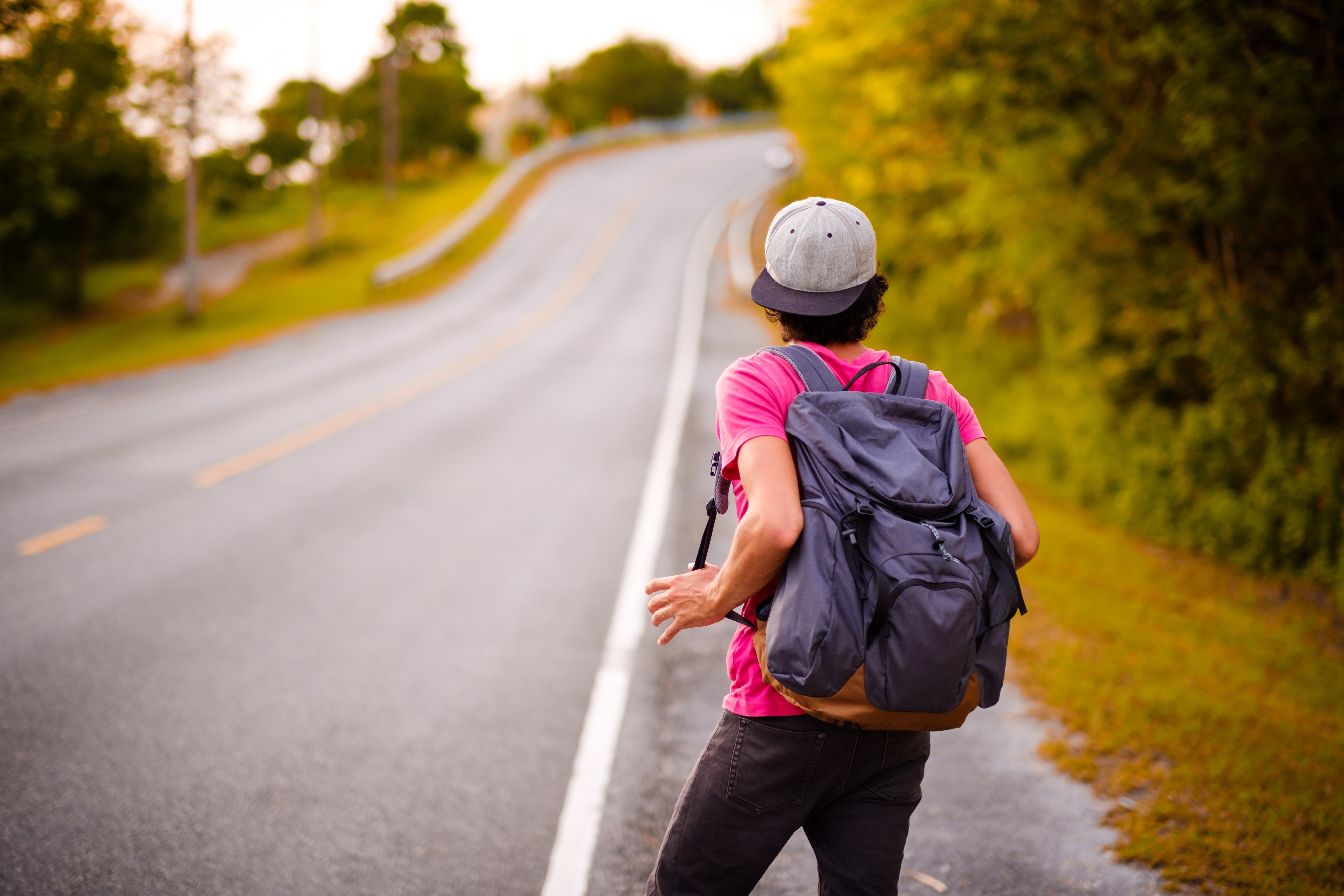 The back of a young man in a pink T-shirt with a blue rucksack and cap, standing at the side of an empty road waiting to be picked up