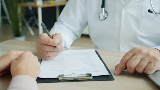 Médico con bata y fonendoscopio rellenando un formulario junto a un paciente, como el cuestionario de salud del seguro médico