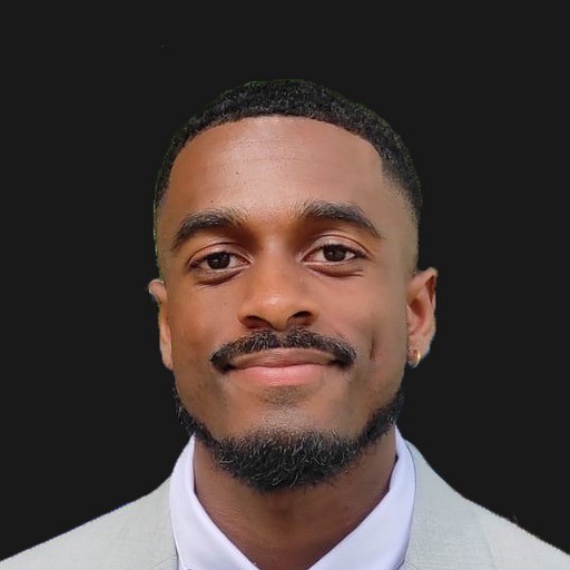 Man with short hair and beard wearing a dark blazer and black shirt against a plain background.
