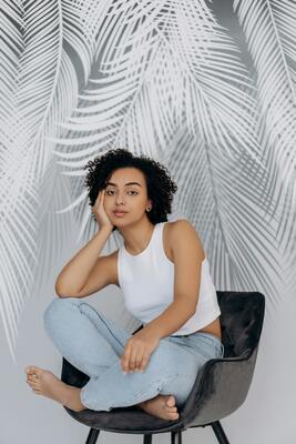 A full-length studio portrait of a woman with curly hair, dressed in a casual white top and light blue trousers, sitting comfortably in a modern black chair against a backdrop of large, decorative palm fronds.