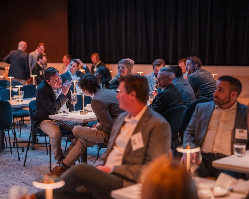 Event attendees talking at small tables with drinks and lamps in a dimly lit room