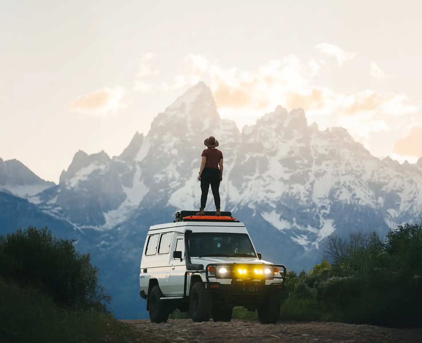 A woman standing on top of an SUV looking out at a jagged mountain peak.