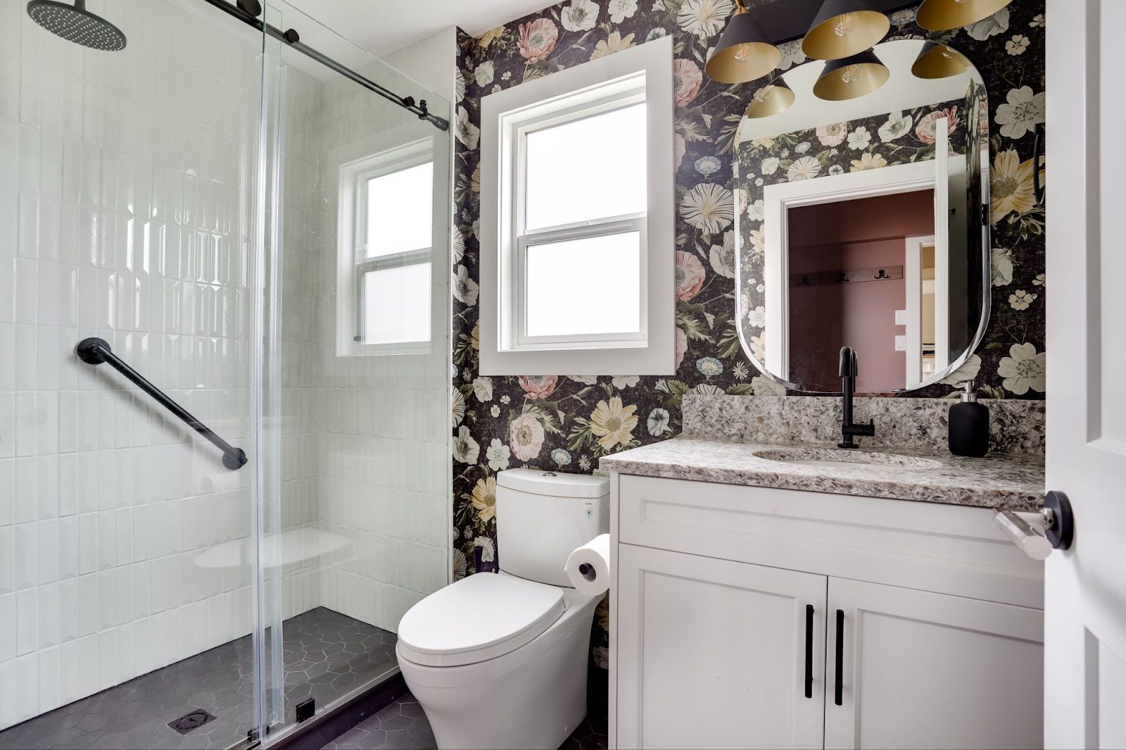 Modern bathroom with a floral accent wall, white vanity with a stone countertop, black fixtures, and a glass-enclosed shower with white tile and a black grab bar.