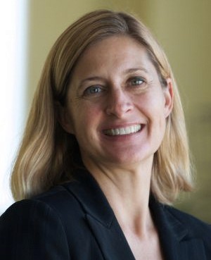 A smiling woman with blonde hair, wearing a dark blazer, stands in a well-lit indoor setting.