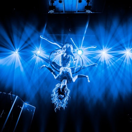 Dua akrobat tampil di atas lingkaran udara, tergantung di bawah sorotan lampu panggung biru terang.