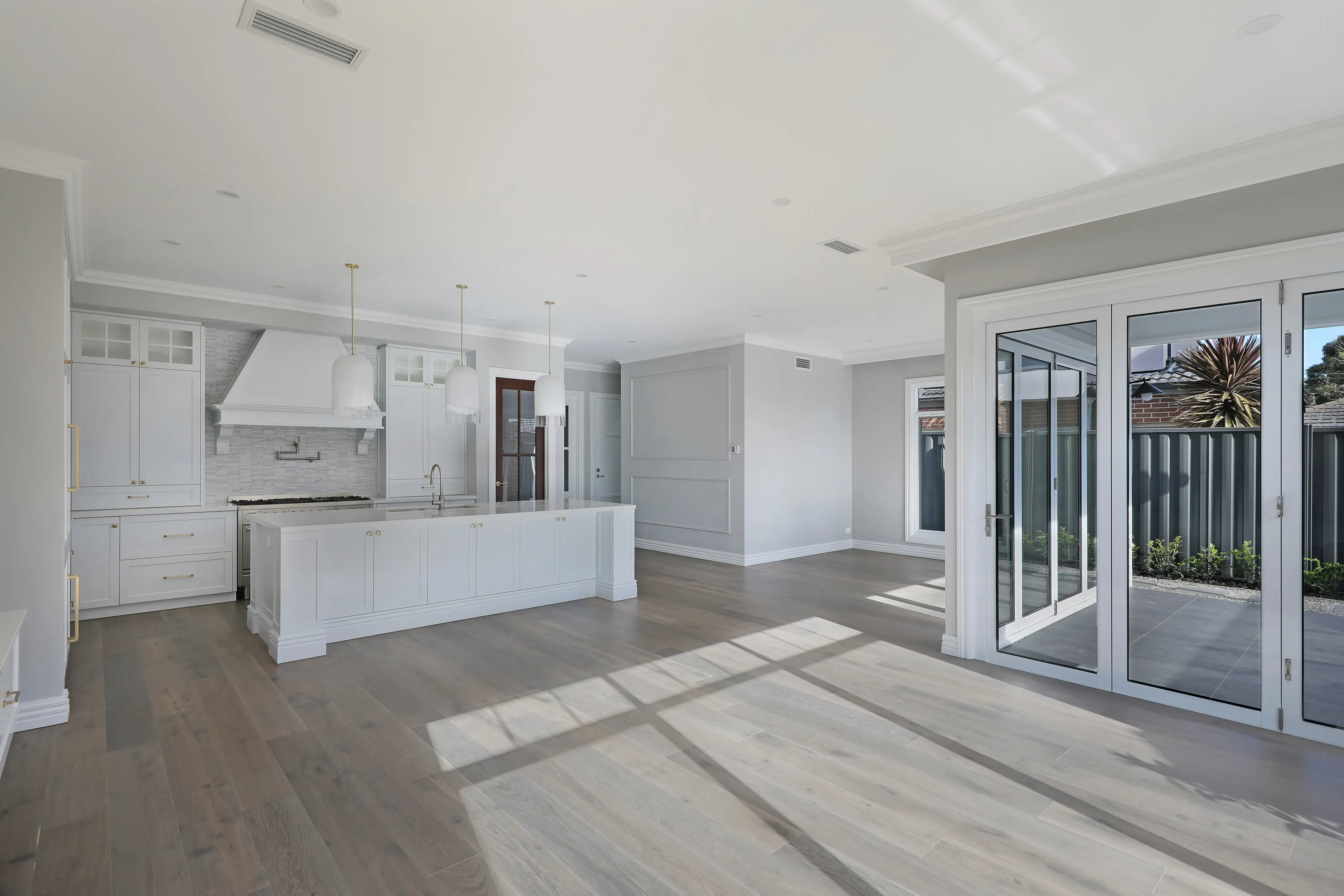 Open-plan living and kitchen with grey walls, dark timber floors, a white island with dome pendants and mantle hood, and white-framed bi-fold doors opening to a tiled courtyard