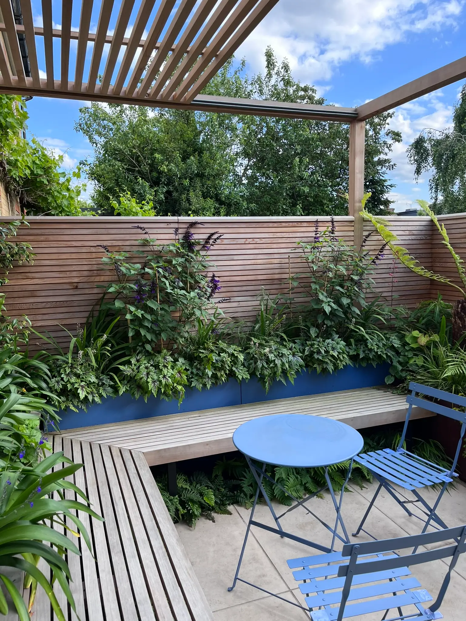 A serene garden patio with a wooden deck, blue table, and lush greenery under a clear sky.