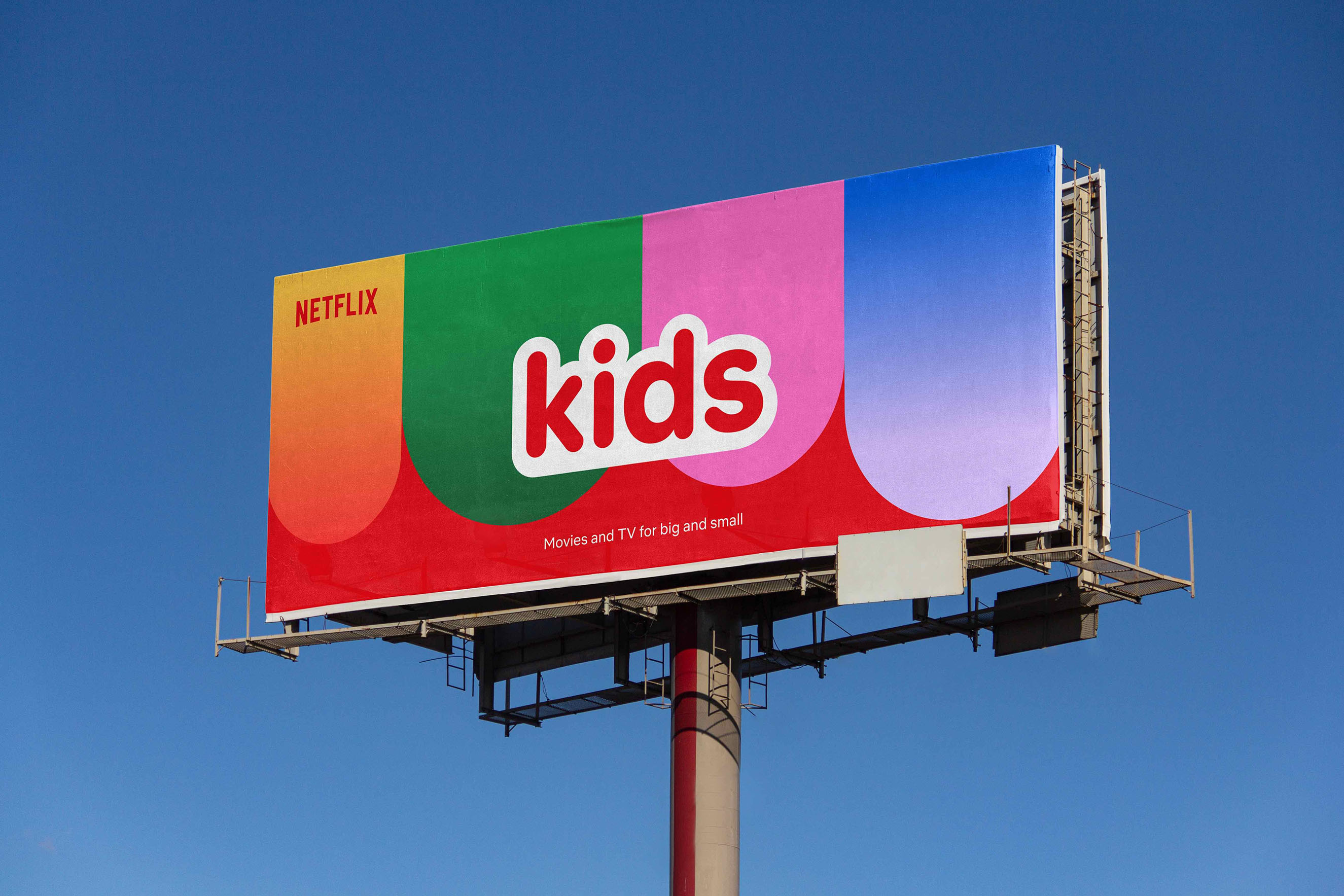 A large outdoor billboard against a clear blue sky featuring the "Netflix Kids" logo over a bold, multi-colored geometric pattern. The tagline "Movies and TV for big and small" illustrates a clean, high-impact brand identity system applied to global out-of-home advertising.