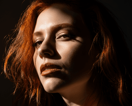 A person with striking red hair and dramatic shadows on their face gazes intensely, creating a moody and artistic portrait in warm lighting.