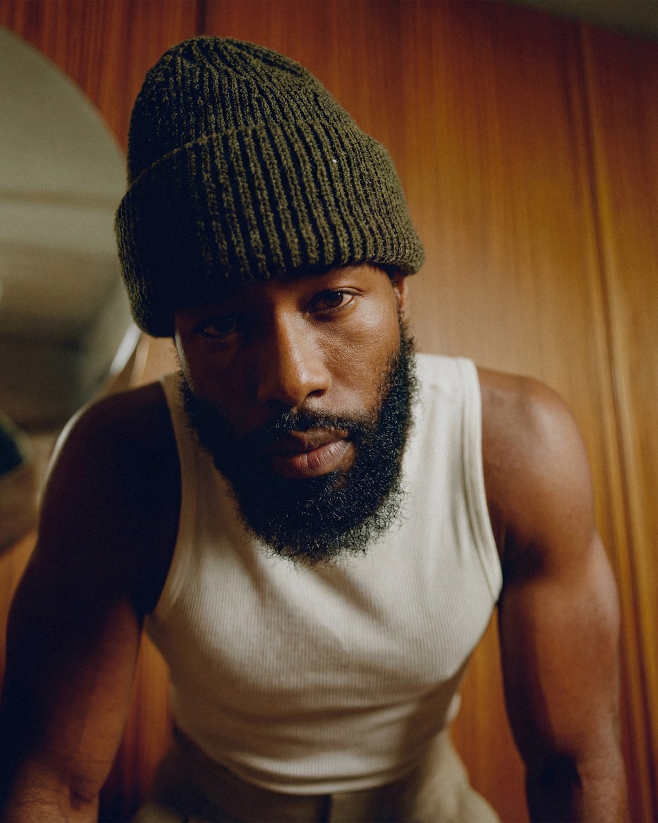 Close-up portrait of a man with a beard wearing a green beanie and white tank top.