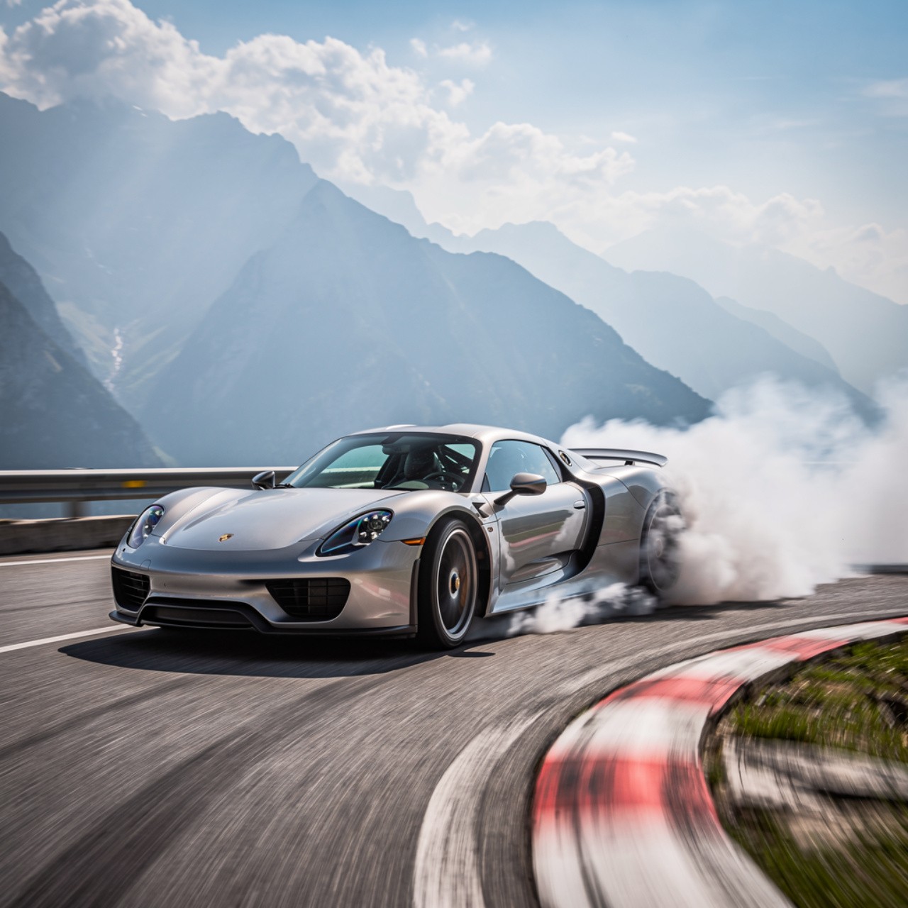 Silver sports car skidding on a racetrack, producing smoke. Majestic mountains and a bright sky in the background convey speed and thrill.