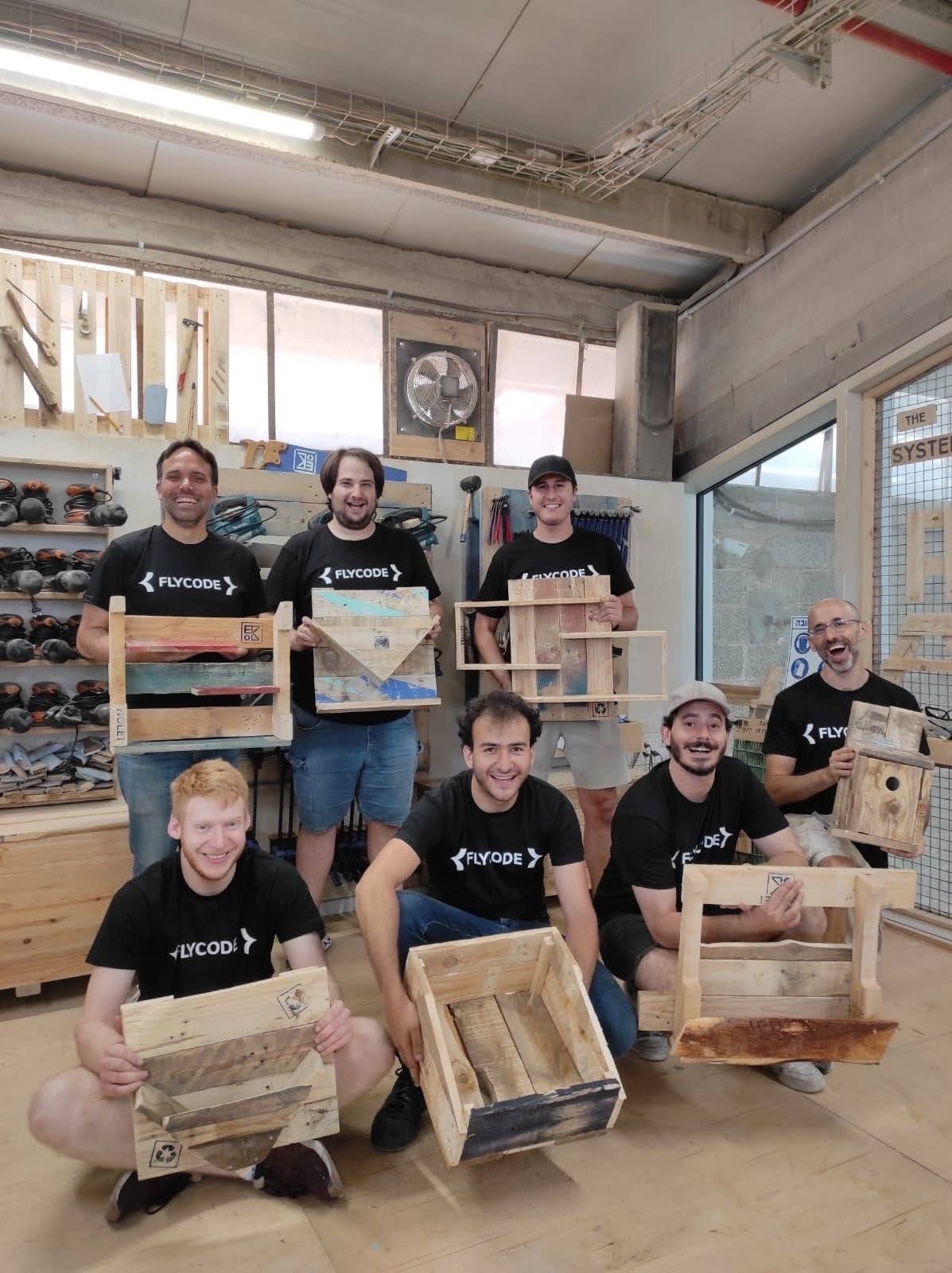 A group of people stands with wooden boxes in a well-lit workshop, all wearing matching black shirts.