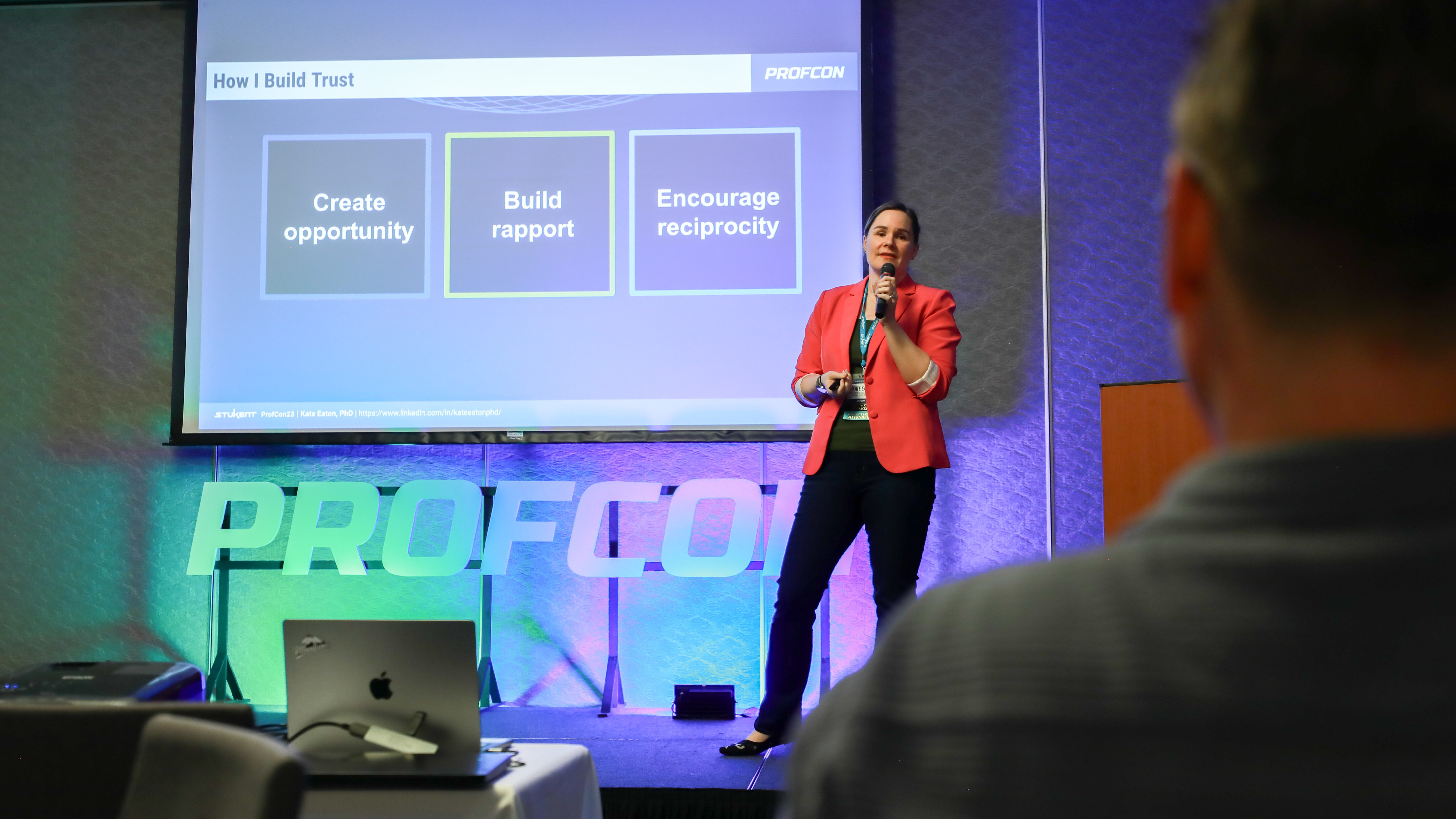 Speaker presents on stage with slide reading: "How I Build Trust – Create opportunity, Build rapport, Encourage reciprocity." PROFCON branding is visible on screen and stage backdrop. Audience member is visible in foreground.
