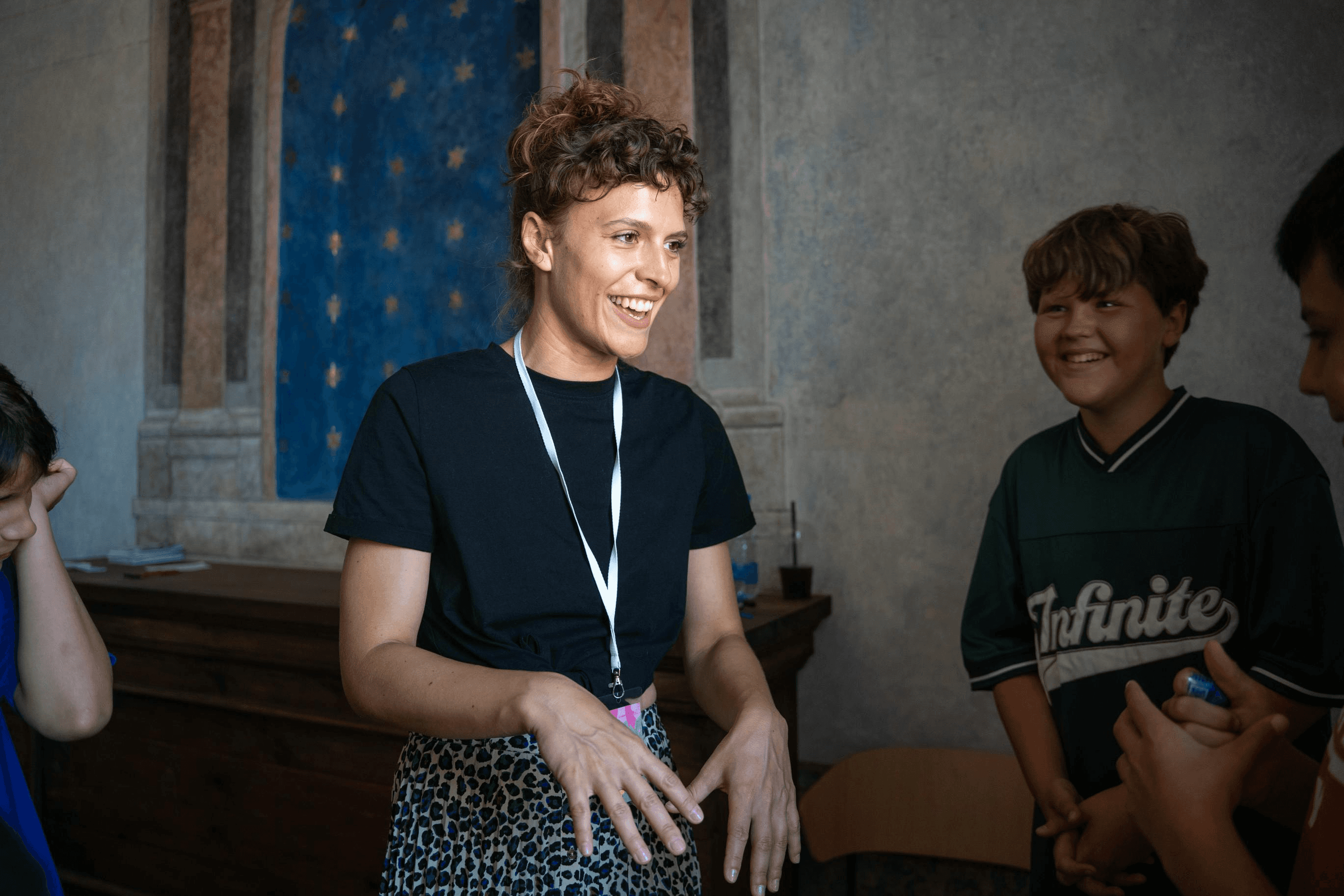 A smiling individual with short curly hair and a lanyard stands animatedly talking with a group of children in a warmly lit room featuring a backdrop of painted stars on the wall.