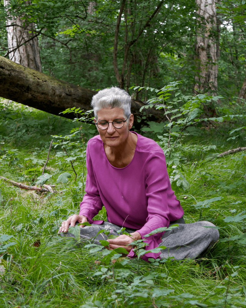 Person mit grauen Haaren und Brille sitzt im Schneidersitz im grünen Wald. Sie trägt ein pinkfarbenes Oberteil und graue Hose und betrachtet achtsam Pflanzen auf dem Waldboden.