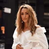 A woman with long, wavy hair wearing a white blouse stands outdoors, looking thoughtfully at the camera.