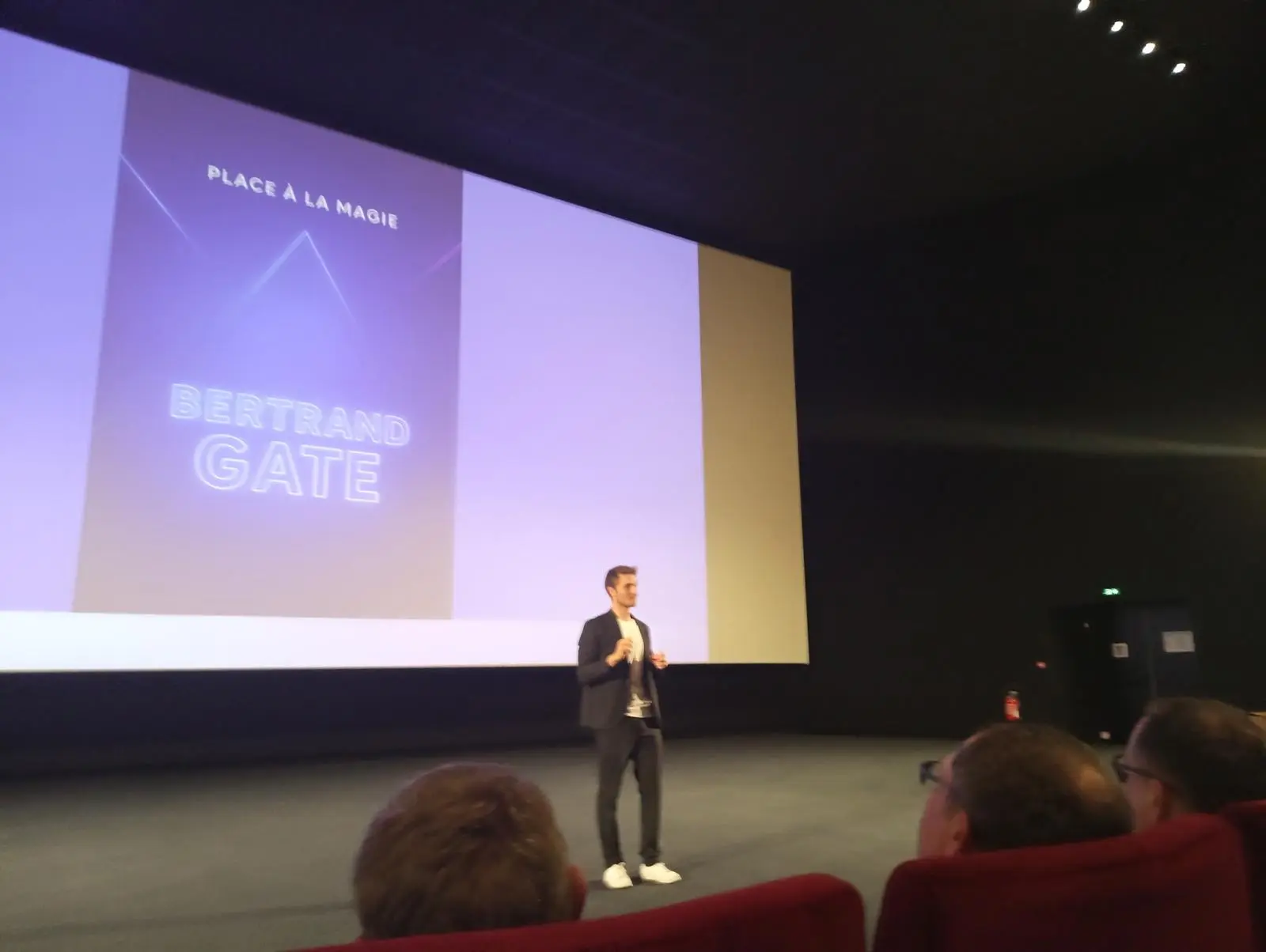 Bertrand Gate, magicien mentaliste, en spectacle de plénière sur scène devant 300 collaborateurs d'IDEMIA lors d'une convention d'entreprise. L'immense écran de projection derrière lui affiche clairement son nom 'BERTRAND GATE' et 'PLACE À LA MAGIE'.